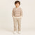 A young boy stands against a plain light beige studio background wearing a cream and brown horizontally striped long-sleeve knit sweater, beige pants, and white sneakers.