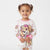 A young girl with brown hair in pigtails wearing a white long-sleeve pajama top with pink trim and a large cartoon dog character print, paired with matching white pajama pants, against a light gray studio background.