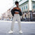 A woman stands on a city street wearing light gray jogger sweatpants, a black sports bra with a white band, a black jacket draped off her shoulders, and white sneakers.