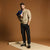 A man stands in a studio wearing a beige crewneck sweatshirt, black cargo jogger pants, and black sneakers, holding a blue plaid shirt under his arm against a warm brown backdrop.