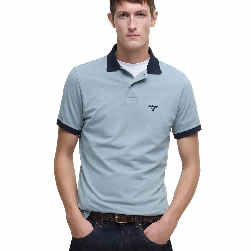 A man wearing a light blue short-sleeve polo shirt with a dark navy collar and sleeve cuffs, paired with dark jeans and a brown belt, against a plain white background.
