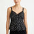 A woman wearing a black camisole top with a small white floral print, lace trim at the neckline, and thin shoulder straps, photographed against a plain light background.