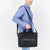 A man in a blue shirt carries a black briefcase-style shoulder bag with a top handle, front zip pocket, and a small red and white stripe detail on the front, shown against a plain white background.