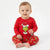 A baby sitting cross-legged wearing a red long-sleeve Grinch pajama top and matching red pajama pants with a green Grinch print, against a plain white background.