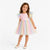 A young girl modeling a pastel tulle dress with a light pink bodice and a layered rainbow tulle skirt, paired with cream Mary Jane shoes, against a plain white studio background.