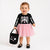 A toddler stands against a plain light background wearing a black long-sleeve top with a white skeleton print and a pink tulle skirt, holding a black tote bag with ghost and heart graphics.