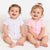 Two smiling babies sitting side by side on a white textured blanket. The baby on the left wears a white short-sleeve bodysuit with a small pink bow pattern, and the baby on the right wears a solid light pink short-sleeve bodysuit.