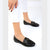 A person wearing black leather loafers with a silver-tone chain detail across the vamp, paired with white cuffed pants, shown against a clean white studio background.