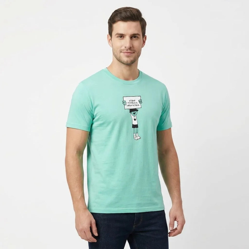 A man wearing a mint green crewneck t-shirt with a cartoon graphic on the chest, paired with dark jeans, standing against a plain white studio background.