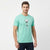 A man wearing a mint green crewneck t-shirt with a cartoon graphic on the chest, paired with dark jeans, standing against a plain white studio background.
