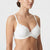 A woman wearing a white wireless bra with delicate lace detailing on the cups, shown from the chest up against a light gray studio background.