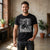 A man wearing a black graphic t-shirt with a white horse design and text, standing in a rustic indoor setting with brick walls and potted plants.