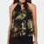 A woman wearing a black halter-neck blouse with a metallic gold floral pattern, featuring a keyhole cutout at the collar and a layered ruffle hem, set against a plain light gray background.