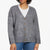 A woman wearing a gray heathered knit cardigan with brown buttons over a white crewneck shirt and blue jeans, shown from the mouth down on a plain white background.