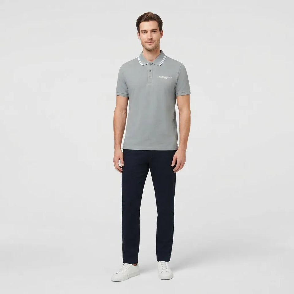 A man modeling a light gray short-sleeve polo shirt with a white embroidered logo on the chest, paired with dark navy pants and white sneakers, against a light gray studio background.