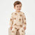 A young child wearing a beige long-sleeve pajama set with a brown teddy bear pattern, sitting on a light-colored chair against a plain white background.