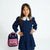 A young girl with long brown hair stands wearing a navy blue long-sleeve dress with a white lace Peter Pan collar and white lace cuffs. She is posing next to a pink and navy blue backpack with the words 'Brains & BEAUTY' on the front pocket, set against a plain white studio background.
