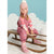 A young girl wearing a pink winter ski suit, matching hat, and boots sits on a wooden sled against a light blue background with white cloud shapes.