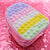 A pastel rainbow pop-it style pouch or small bag with a glossy iridescent pink body and a translucent top panel featuring horizontal rows of bubble textures in lavender, pink, white, yellow, and mint, shown on a shiny pink reflective surface.