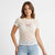 A woman wearing a short-sleeve lace top with a pink bow graphic on the chest, paired with light blue jeans, against a plain white studio background.