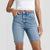 A model wearing light-wash blue denim biker shorts with a raw, frayed hem, paired with a white cropped tank top against a light gray studio background.
