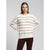 A woman modeling a cream and beige horizontally striped knit sweater with dark brown pants, set against a plain light gray studio background.