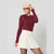 A woman posing in a studio setting wearing a burgundy long-sleeve knit top, an off-white pleated mini skirt, a beige newsboy cap, and black tights.