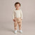 A toddler standing against a plain beige studio background, wearing a cream long-sleeve top and beige patterned jogger pants with white sneakers.