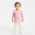 A toddler girl with curly light brown hair stands against a plain white studio background wearing a light pink ribbed short-sleeve top with ruffled edges, white pants, and white shoes.