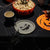 Halloween-themed tabletop scene with a bowl of popcorn, a black mug, a paper plate with broken glass pieces, spider decorations, and an orange jack-o'-lantern decoration on a black tablecloth.