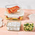 A set of clear glass rectangular food storage containers with white lids, shown filled with food on a pink surface against a white background.