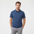 A man wearing a heathered blue short-sleeve polo shirt and light gray trousers, standing against a plain white studio background.