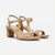 A pair of women's block-heel sandals in a light tan/beige color, featuring a low block heel, double front straps, and an ankle strap with a silver buckle, shown against a plain light gray background.