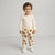 A toddler standing against a light gray studio background, wearing a cream long-sleeve top and matching cream pants with a brown teddy bear print, plus white sneakers.