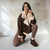 A woman crouches in a studio wearing a brown shearling-lined jacket with cream trim, matching ribbed brown leggings, and brown platform ankle boots against a textured plaster wall.