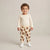 A toddler standing against a light beige studio background, wearing a cream long-sleeve top and matching cream pants with a brown teddy bear print, paired with white sneakers.