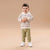 A toddler standing against a solid beige studio background, wearing a cream and brown horizontally striped long-sleeve top with a white collar, olive green ribbed pants, and white sneakers.