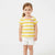 A young girl with light brown hair in a ponytail stands against a plain white studio background, wearing a yellow and white horizontally striped short-sleeve t-shirt with a pink collar and a small colorful graphic on the chest, paired with white shorts.