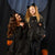 A woman and a young girl wearing matching black cat-themed Halloween costumes, standing in front of a dark curtain backdrop with pumpkins at the bottom.