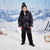 A young boy wearing a black and gray camouflage winter ski suit, black beanie with a pom-pom, and black ski boots, standing in a snowy landscape with a blue and white mountain backdrop. A blue and white striped beach chair is visible on the right.
