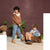 Two children modeling coordinated outfits in a styled indoor scene with a textured mauve wall, plants, and a small wooden cart. One child stands wearing a brown zip-up hoodie over a cream shirt and light blue joggers, while the other sits in a wooden cart wearing a light blue hoodie and tan pants.