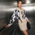 A woman posing in a modern metallic hallway wearing a white long-sleeve blouse with a black floral pattern, a beige knit scarf, a beige mini skirt, and holding a black textured clutch.