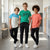 Three boys standing in a school hallway wearing matching athletic sets: light blue and white graphic T-shirts, a green graphic T-shirt, and coral pink graphic T-shirts, paired with black jogger pants and white sneakers.