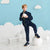 A young boy stands in a studio setting wearing a navy blue tracksuit with a bright lime green drawstring. He is balancing on a white spherical platform, holding a white ball, with a small white bench and cloud cutouts against a light blue background.