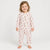A smiling toddler stands barefoot against a plain light gray studio background, wearing a long-sleeve footless one-piece pajama or romper with a front zipper and an allover small teddy bear and floral print.