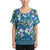 A woman wearing a short-sleeve floral top with cold-shoulder cutouts. The top features a vibrant pattern of blue, teal, green, and white flowers with a striped hem, set against a plain white background.