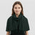 A woman with shoulder-length brown hair wearing a dark forest green textured knit scarf with fringe, styled over a black top against a light gray studio background.