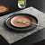 A styled tabletop setting with a dark charcoal gray plate holding a speckled terracotta plate and a gold-toned fork, placed on a gray woven placemat over a dark table. A glass of water with a lemon slice and stacked matching plates appear in the background.