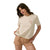 A woman modeling a cream-colored short-sleeve t-shirt with a small pink embroidered graphic on the left chest, paired with beige shorts, against a solid white background.