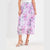 A model wearing a high-waisted midi skirt with a pink and purple watercolor floral print on a white base, paired with beige strappy heeled sandals against a plain white studio background.